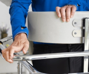 A person in a blue shirt leans on a walker, gripping the handles tightly. The scene conveys determination and support in a healthcare setting.