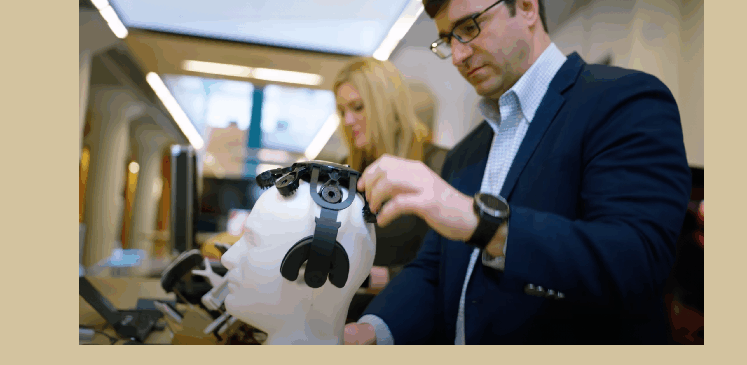 Man in suit adjusts headgear on a mannequin in a tech lab. A woman in the background focuses on equipment. The scene is focused and professional.