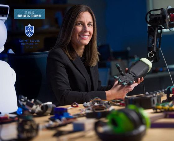 A woman in a black blazer smiles while holding a robotic hand in a workshop. The desk is cluttered with electronic parts. Logos of St. Louis Business Journal and Saint Louis University are in the background.