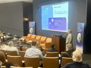 A speaker presents in a modern auditorium at the BioSTL Global Health Innovation Summit. Attendees sit attentively, with a vibrant projection screen behind.